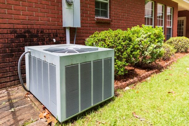 Older style air conditioner system next to home, brick and bushes with clean yard.